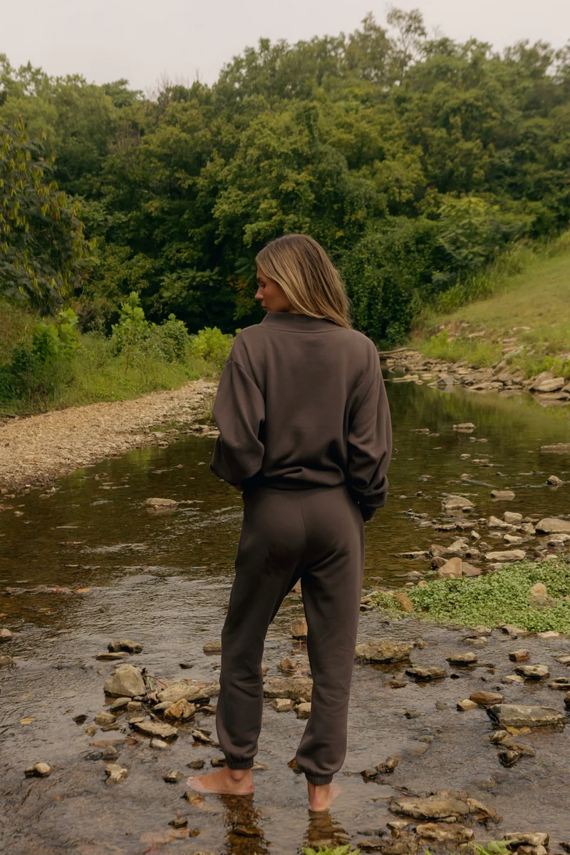 A woman stands barefoot in a shallow rocky stream, viewed from behind, wearing a dark brown matching lounge set consisting of a long-sleeve top and sweatpants, with a lush green forest in the background.