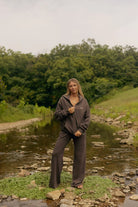 A woman wearing a dark brown zip-up hoodie and matching wide-leg sweatpants stands on a rocky bank next to a stream, with a lush green forest in the background.