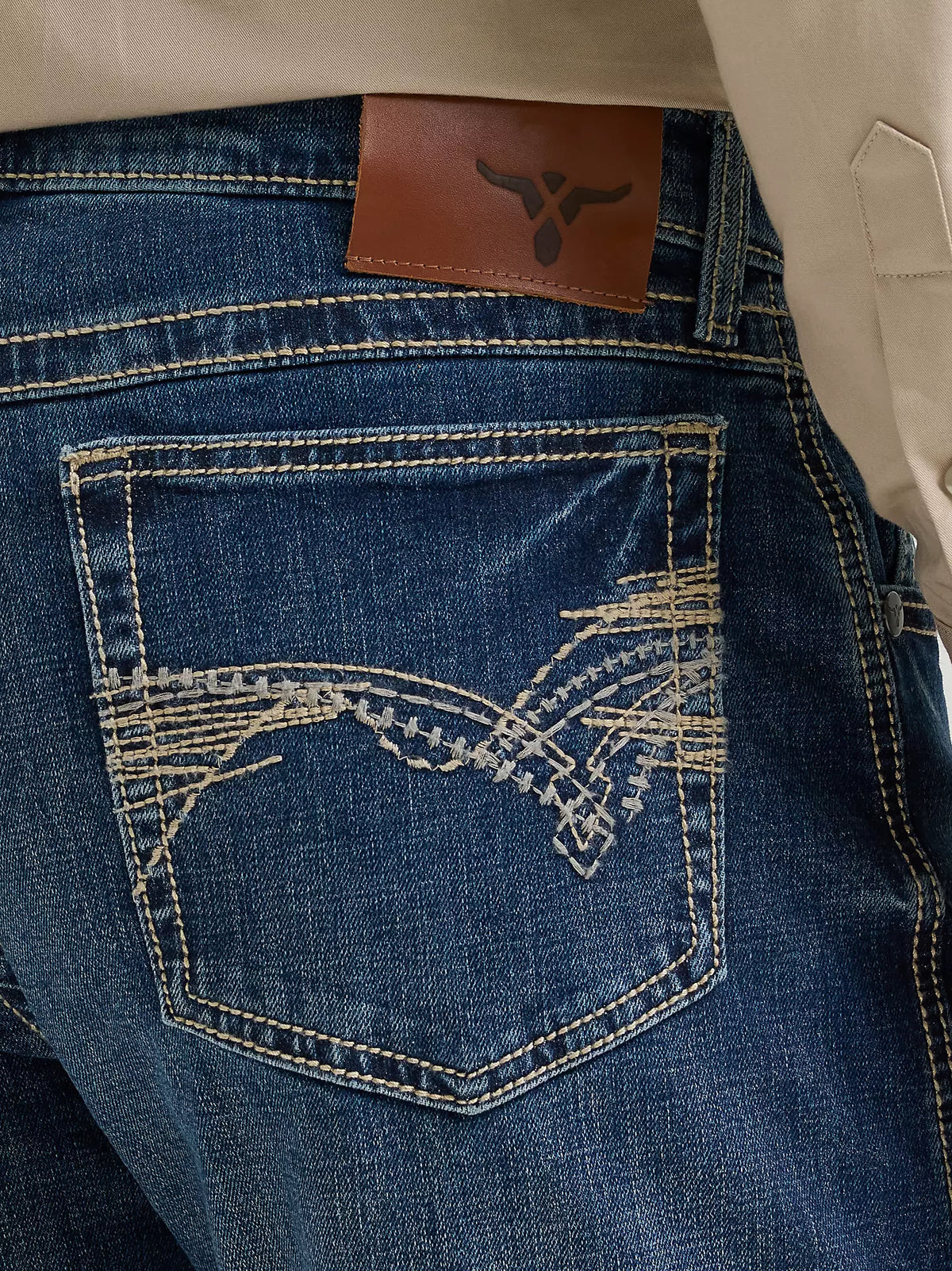 Close-up of the back pocket of blue denim jeans with decorative white embroidery, a brown leather patch, and contrast stitching, shown with a beige shirt at the top.
