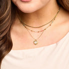 Phoebe colorful gold ball necklace worn between a rope chain and a heart pendant necklace with a white top.