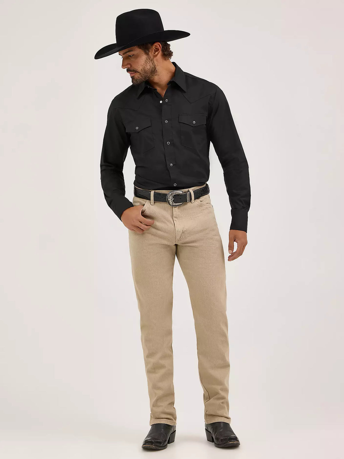 Man wearing a black cowboy hat, black shirt, and beige pants on a white background