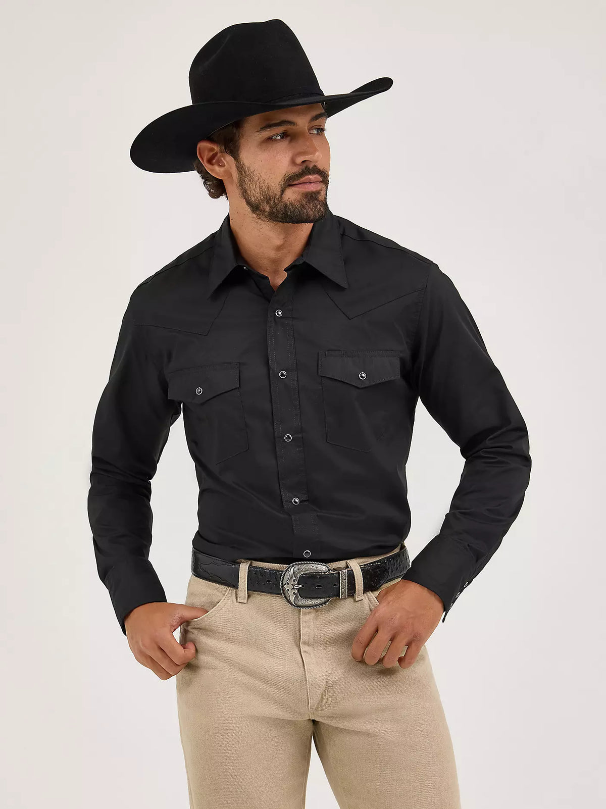 Man wearing a black cowboy hat, black shirt, and beige pants on a white background