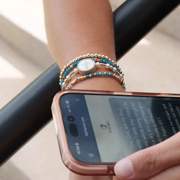 Tap To Pray bracelet worn in a beaded bracelet stack beside a smartphone displaying a Bible verse.
