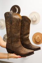 Brown cowboy boots held by a person with decorative hats in the background. Tecovas women’s cowboy boots