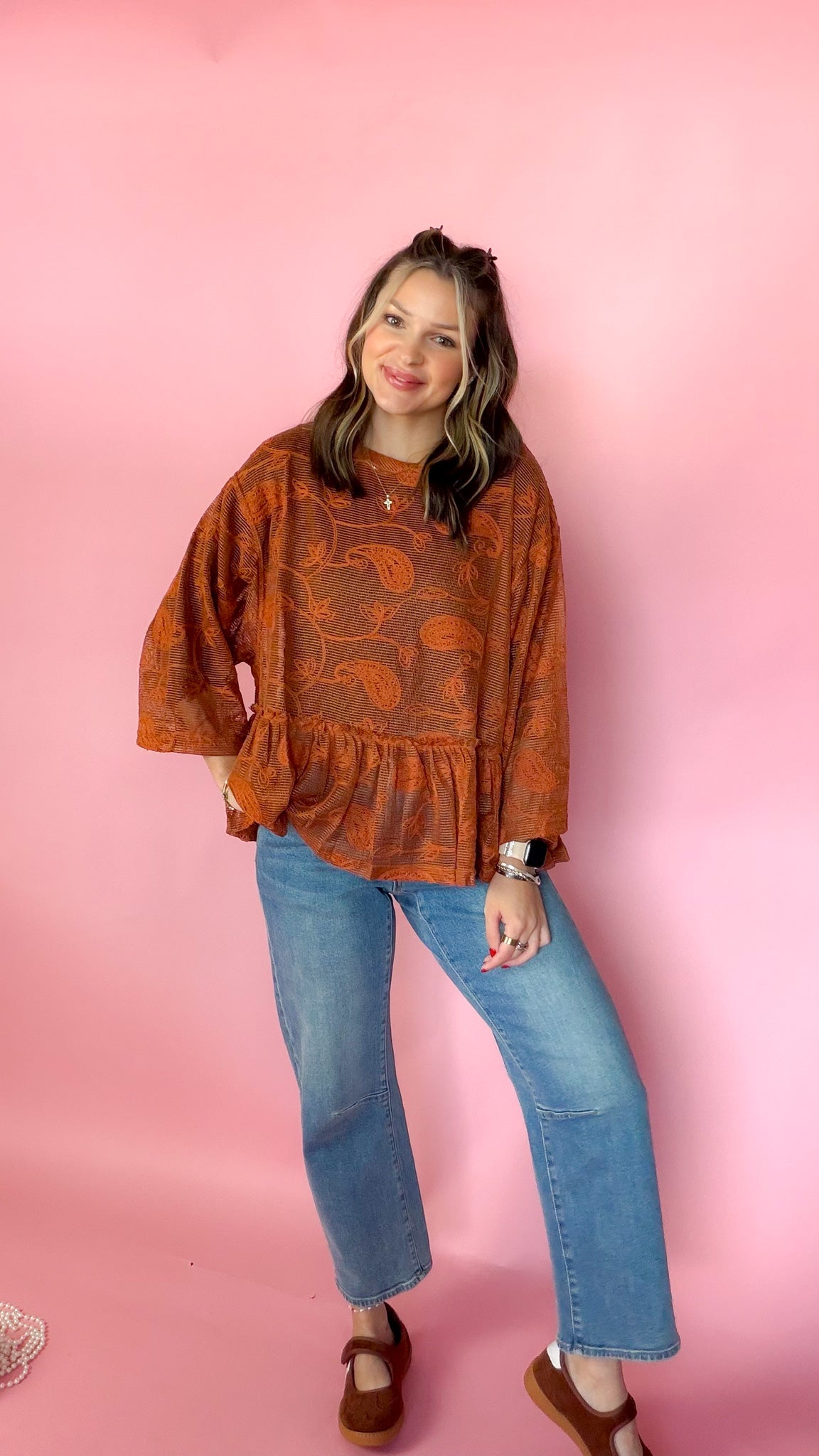 A woman modeling a rust-orange patterned long-sleeve blouse with a ruffled hem, paired with blue jeans and brown shoes, standing against a solid pink background.