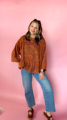 A woman modeling a rust-orange patterned long-sleeve blouse with a ruffled hem, paired with blue jeans and brown shoes, standing against a solid pink background.
