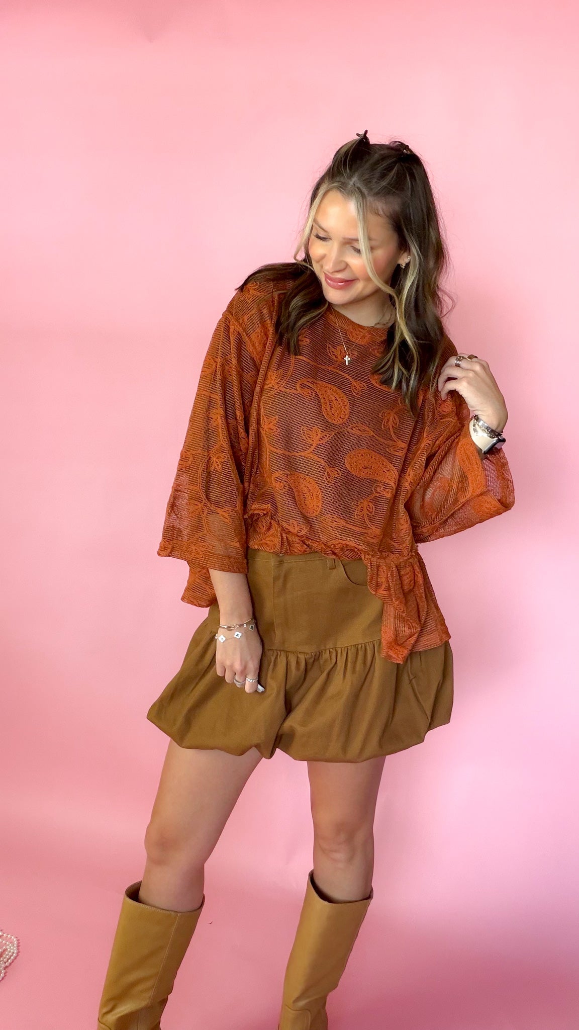 A woman modeling a rust-orange patterned long-sleeve blouse with a ruffled hem, paired with brown skirt and brown boots, standing against a solid pink background.