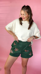 woman standing against pink background wearing cream sweater paired with green mallard shorts