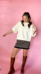 woman standing against pink background wearing cream sweater paired with black plaid skirt and knee high boots. 