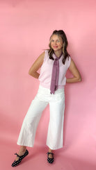 A woman posing in a sleeveless light pink top, white wide-leg pants, a purple striped necktie, and black polka-dot shoes against a solid pink background.