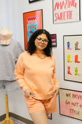 Woman in a green sweater and checkered pants standing in a room with pink couch, decorative pillows, and wall art.
