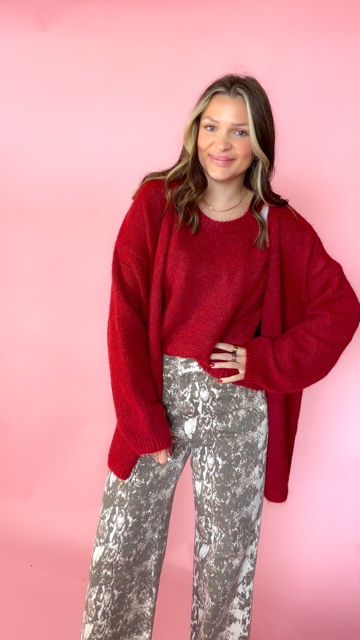 woman wearing a red knit tank with a matching red cardigan against a pink background. Paired with snake print pants