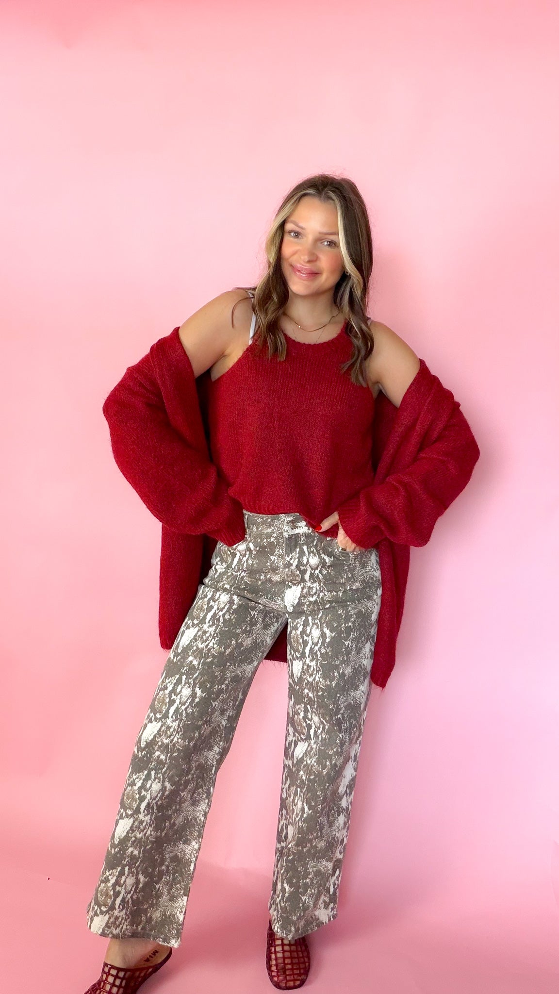 woman wearing a red knit tank with a matching red cardigan against a pink background. Paired with snake print pants