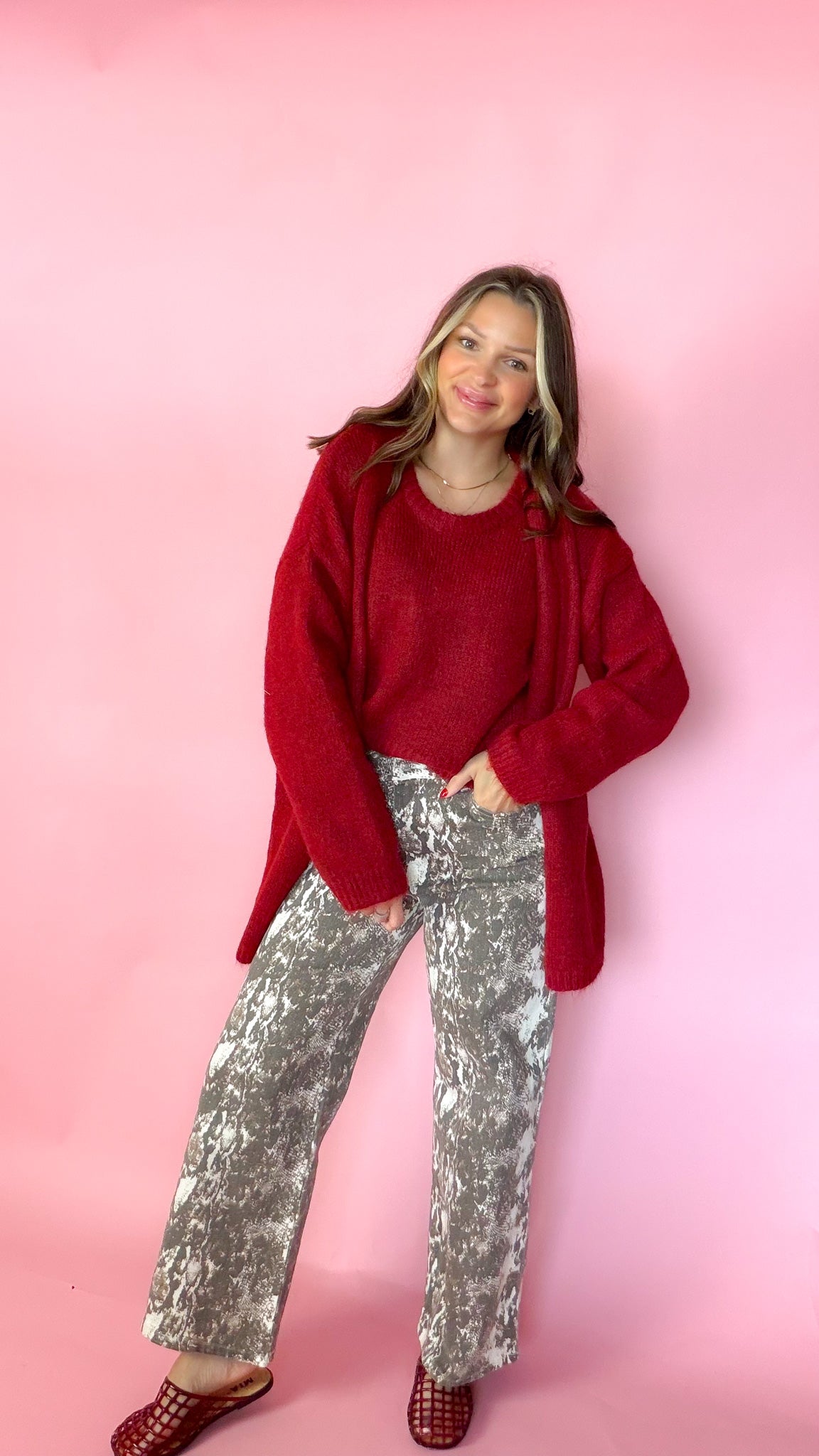 woman wearing a red knit tank with a matching red cardigan against a pink background. Paired with snake print pants