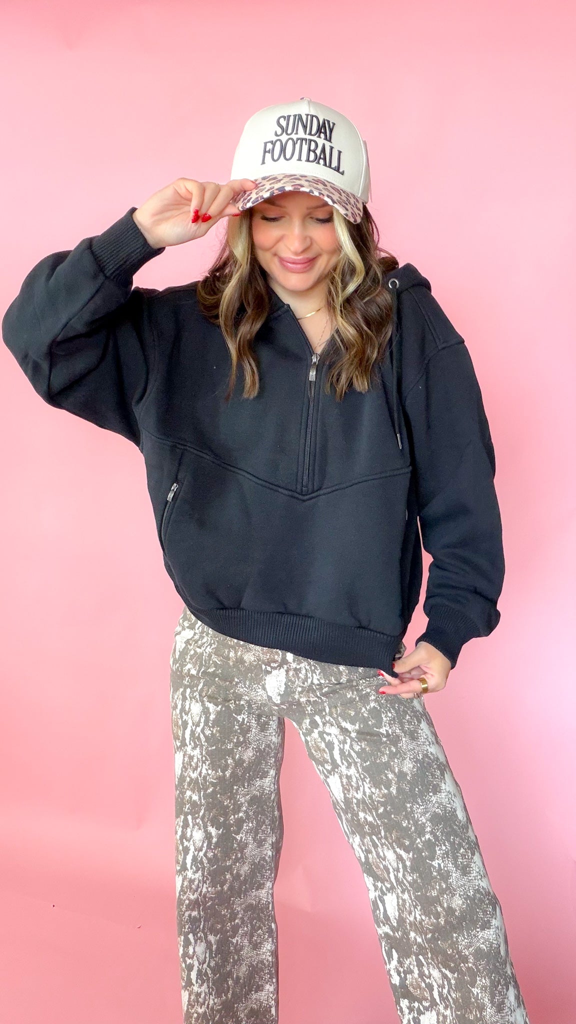woman picturing black half zip pullover with leopard trucker hat and snake print jeans.