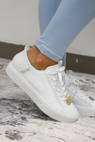White sneakers with a gold logo worn with light blue pants on a marble floor.