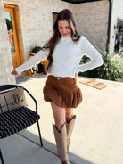 Woman wearing a white sweater and brown shorts with beige cowboy boots, standing on a patio.