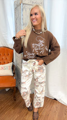 A person wearing a brown sweatshirt with a cowboy graphic and cream pants featuring a brown and gray horse print, standing in a room with a wooden floor, white curtain backdrop, and an orange chair.