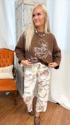 A woman modeling a brown crewneck sweater with a graphic and the words 'Howdy Cowgirl!' paired with cream pants featuring a cowboy and horse pattern, standing in front of a white curtain backdrop.