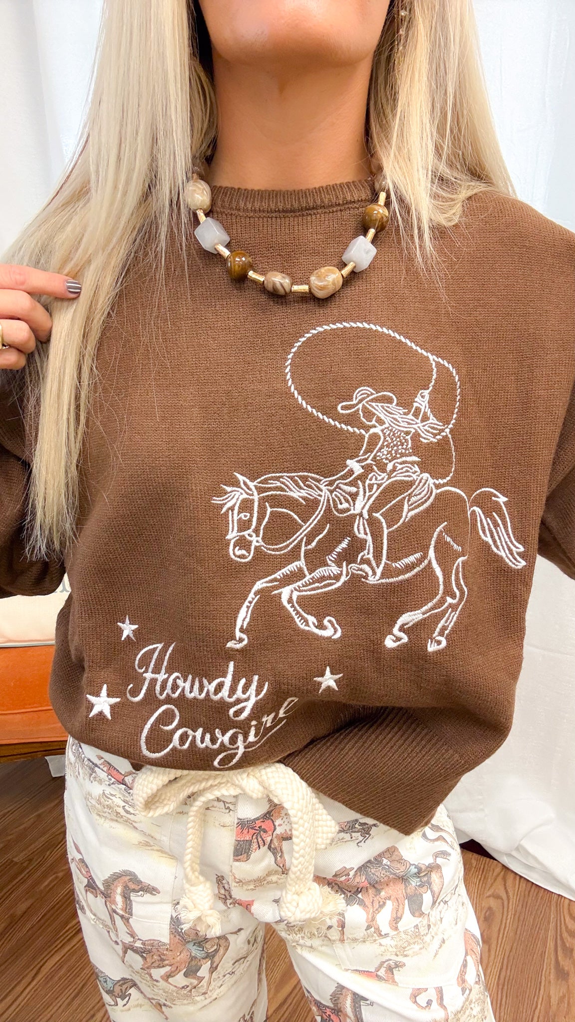 A woman modeling a brown crewneck sweater with a graphic and the words 'Howdy Cowgirl!' paired with cream pants featuring a cowboy and horse pattern, standing in front of a white curtain backdrop.