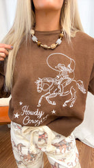 A woman modeling a brown crewneck sweater with a graphic and the words 'Howdy Cowgirl!' paired with cream pants featuring a cowboy and horse pattern, standing in front of a white curtain backdrop.