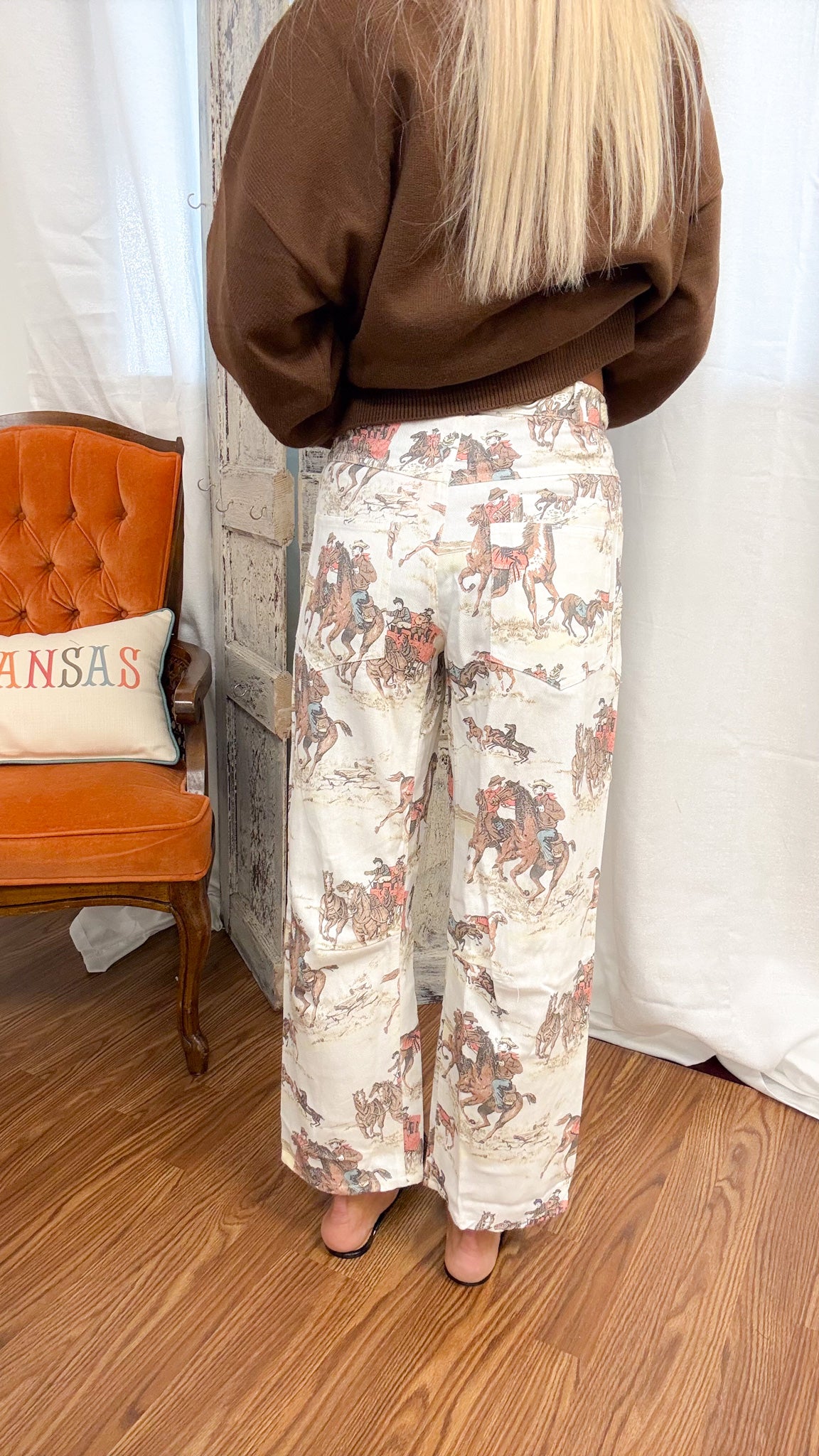 A person shown from the back wearing wide-leg pants with a detailed western-style horse and rider print, paired with a brown sweatshirt. The setting is indoors with a wooden floor, white curtain backdrop, and an orange tufted chair on the left.