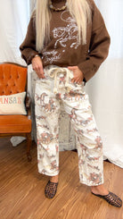 A person wearing a brown sweatshirt with a cowboy graphic and cream pants featuring a brown and gray horse print, standing in a room with a wooden floor, white curtain backdrop, and an orange chair.