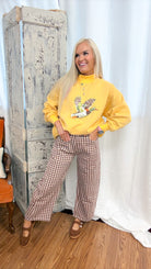 A woman posing in a yellow mock-neck sweatshirt with a duck graphic and brown-and-white gingham wide-leg pants, standing in front of a rustic white curtain and a distressed wooden cabinet.
