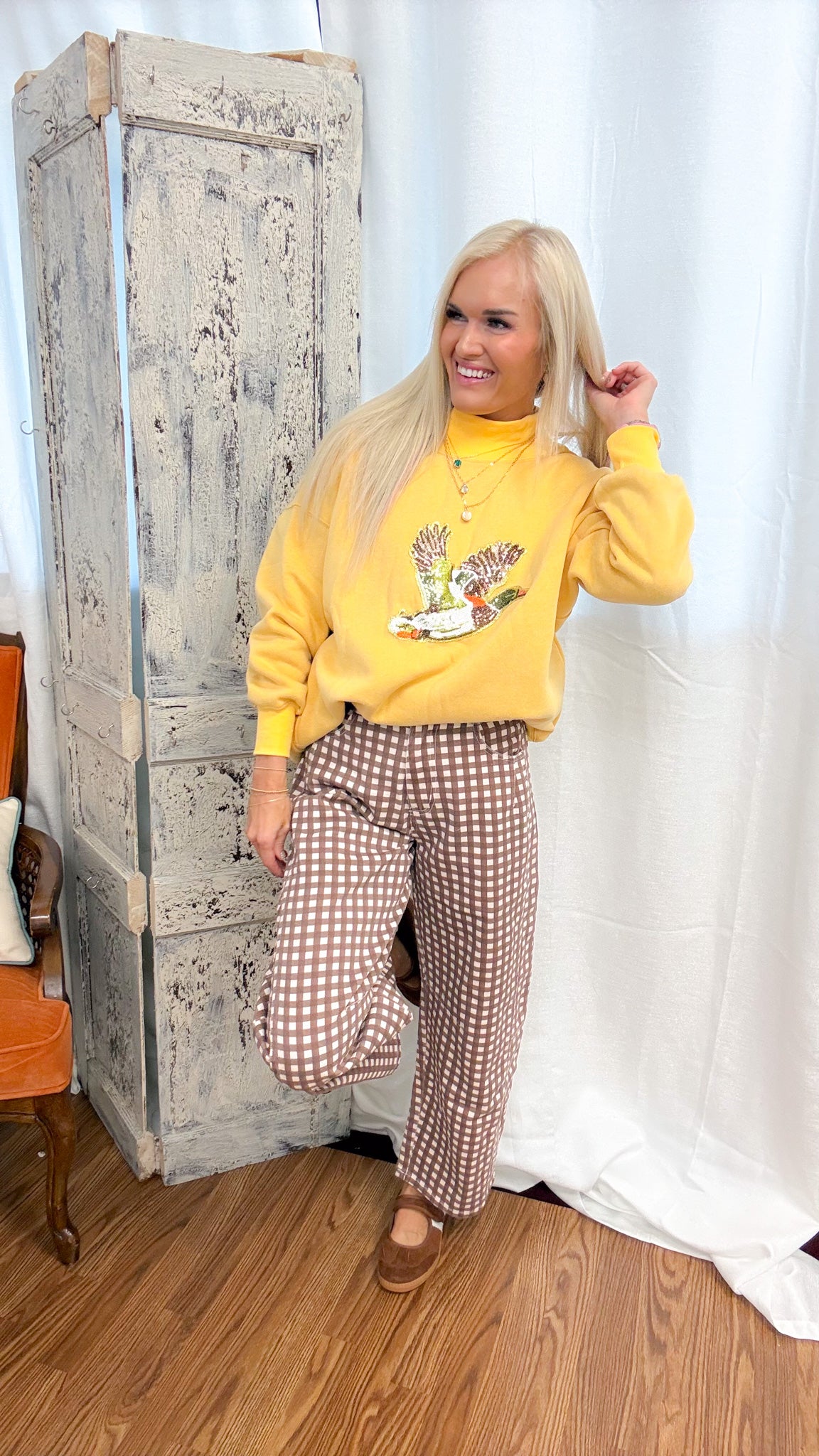 A woman posing in a yellow mock-neck sweatshirt with a duck graphic and brown-and-white gingham wide-leg pants, standing in front of a rustic white curtain and a distressed wooden cabinet.