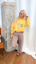 A woman posing in a yellow mock-neck sweatshirt with a duck graphic and brown-and-white gingham wide-leg pants, standing in front of a rustic white curtain and a distressed wooden cabinet.