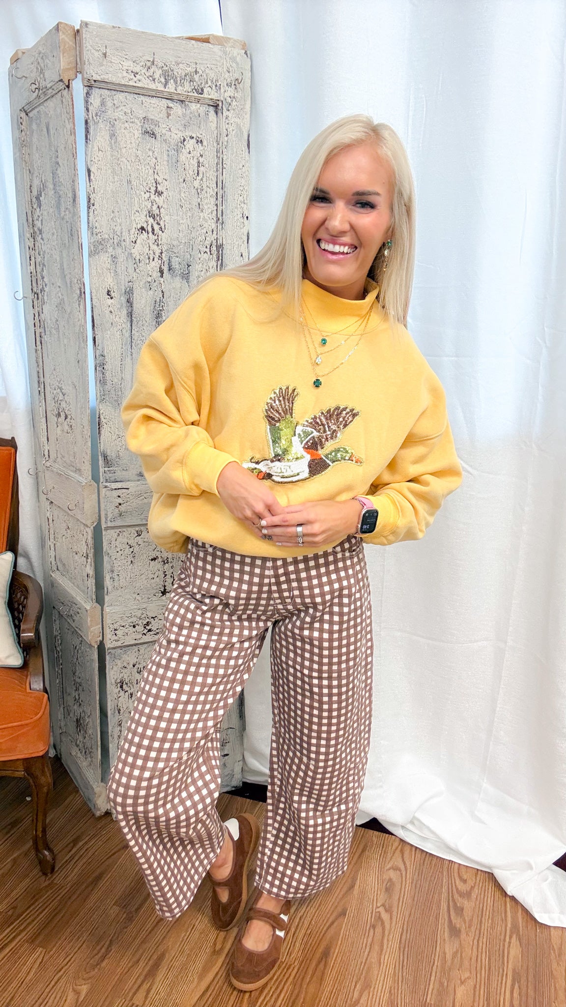A woman posing in a cozy indoor setting, wearing a mustard yellow sweatshirt with a mallard graphic and brown-and-white gingham wide-leg pants.