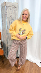 A woman posing in a cozy indoor setting, wearing a mustard yellow sweatshirt with a mallard graphic and brown-and-white gingham wide-leg pants.