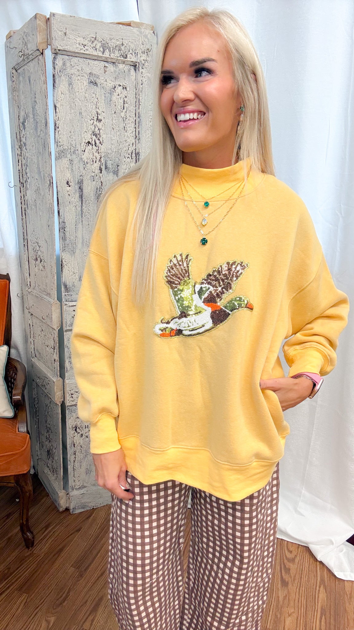A woman posing in a yellow mock-neck sweatshirt with a duck graphic and brown-and-white gingham wide-leg pants, standing in front of a rustic white curtain and a distressed wooden cabinet.