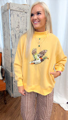 A woman posing in a yellow mock-neck sweatshirt with a duck graphic and brown-and-white gingham wide-leg pants, standing in front of a rustic white curtain and a distressed wooden cabinet.