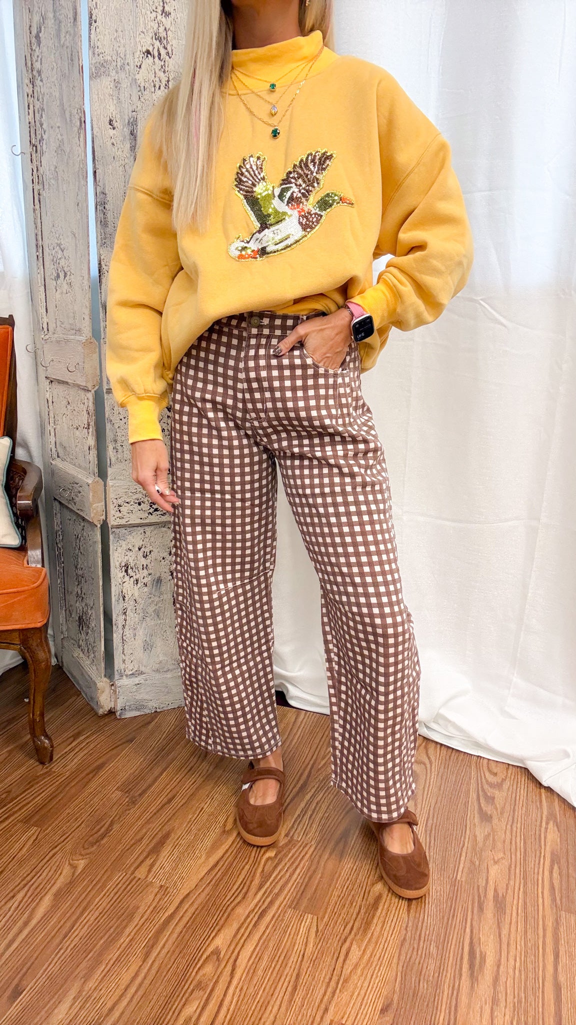 A person wearing a mustard yellow sweatshirt with a duck graphic and brown-and-white gingham wide-leg pants, standing on a wood floor in front of a white curtain.