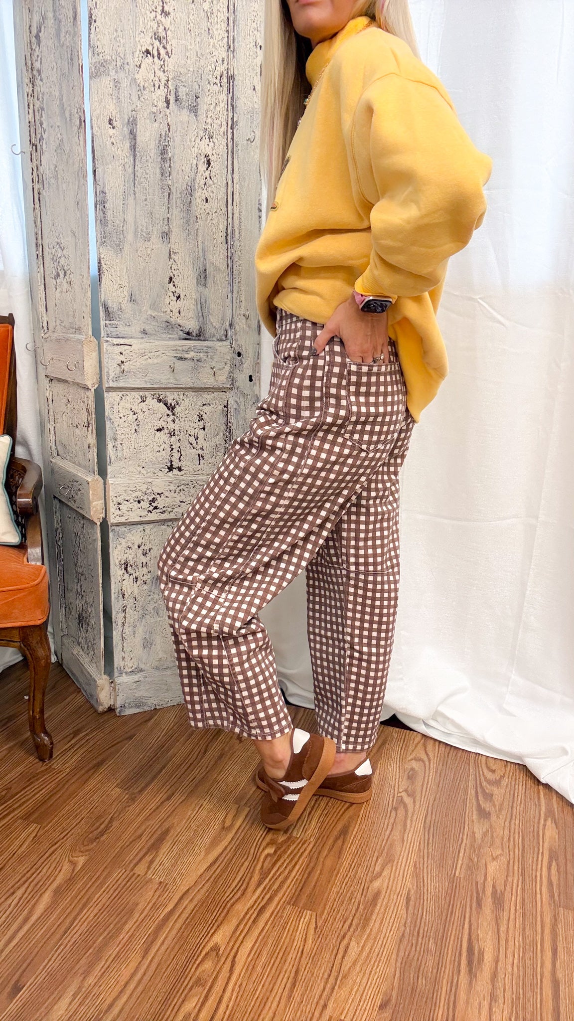 A person wearing brown and white gingham wide-leg cropped pants, a yellow sweatshirt, and brown wedge sandals, standing in a room with a rustic wooden door, white curtain, and wood floor.