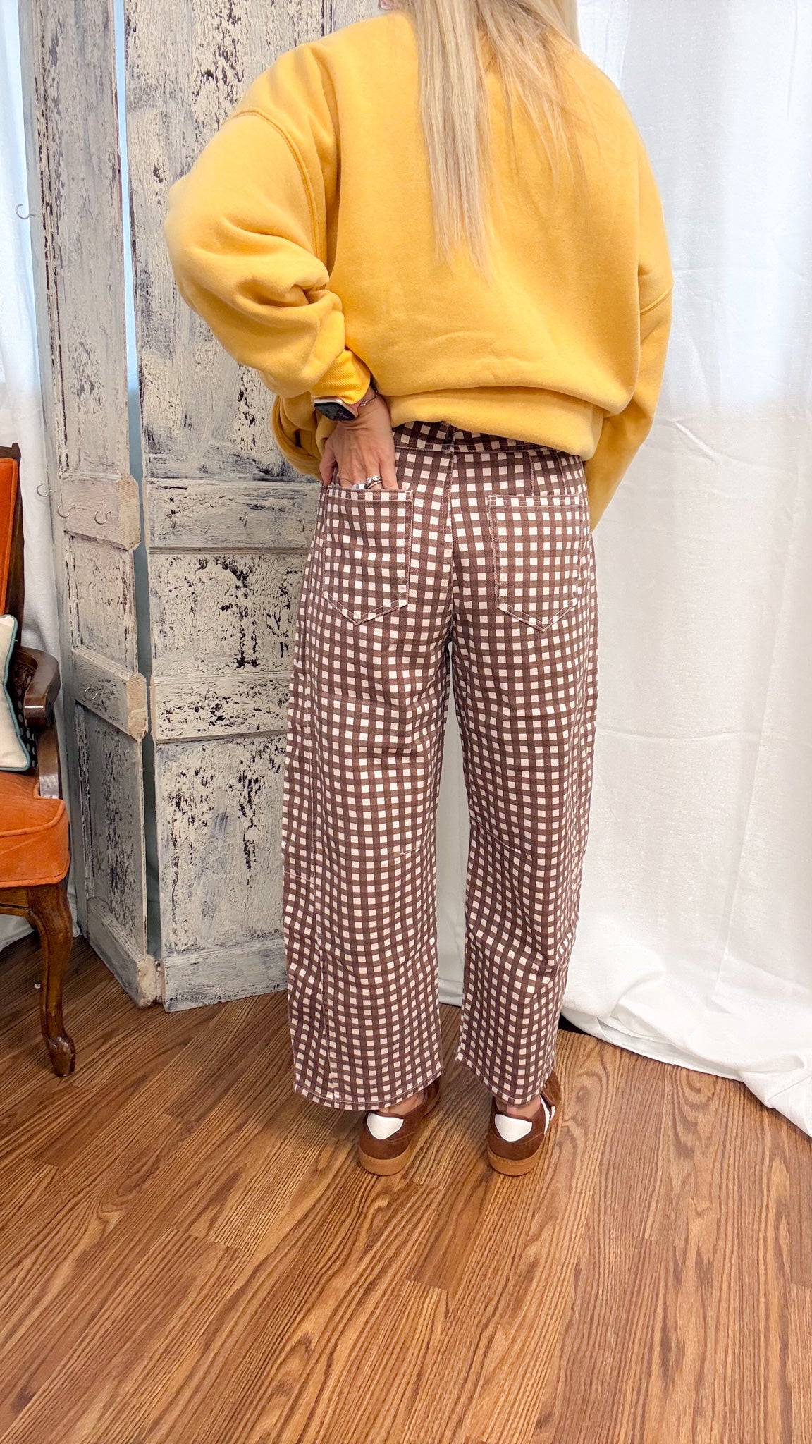 A person shown from the back wearing brown and white gingham wide-leg pants, a yellow sweatshirt, and brown shoes in an indoor room with wood floors and a rustic wall.