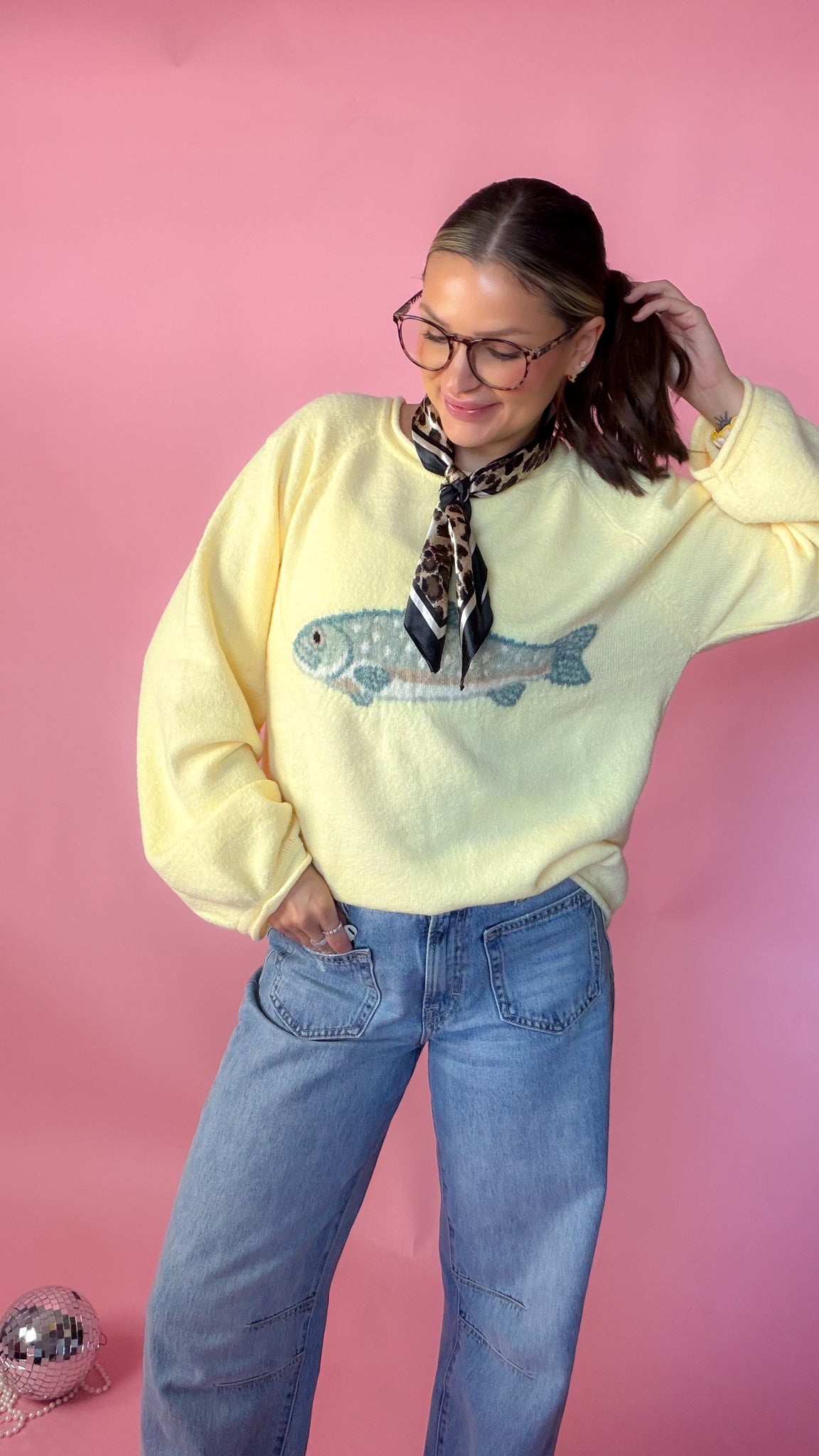 woman standing against pink background wearing yellow fish sweater paired with leopard neck scarf