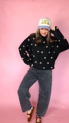 At A Glance black polka dot sweater worn with gray jeans and a trucker hat, playful pose