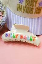 Dilly Dally Hair Claw Clip in cream with rainbow letters, shown in front of a trucker hat