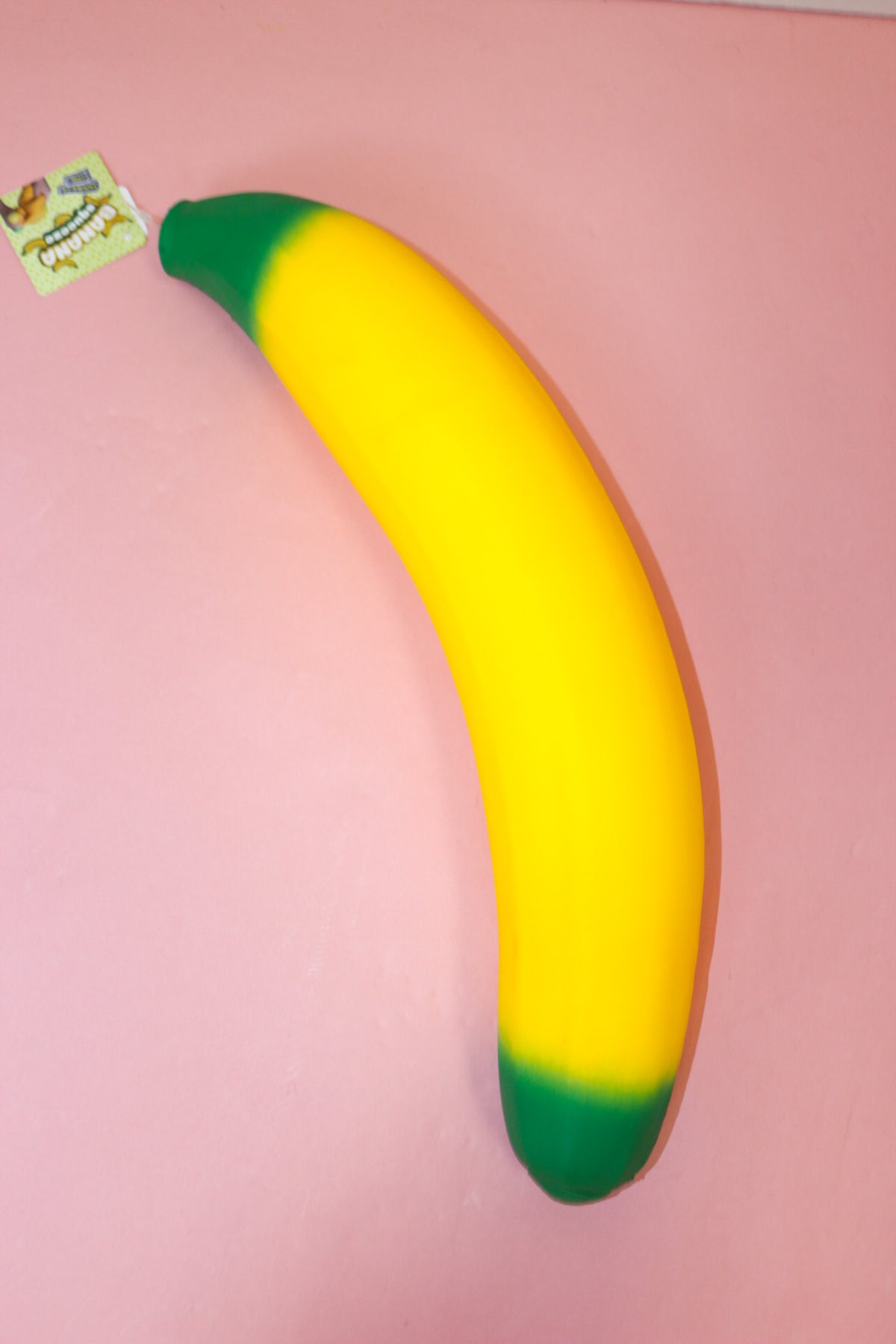 Jumbo yellow banana squishy with green ends and a curved shape lying on a pink surface.