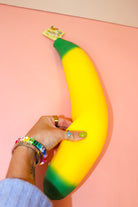 Hand squeezing the middle of a jumbo yellow banana squishy with green ends on a pink surface.
