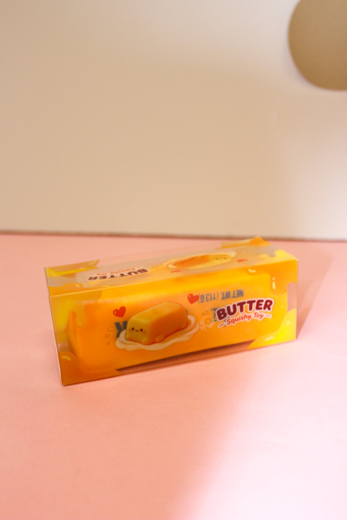 Butter squishy toy in a yellow rectangular box with butter-themed graphics displayed on a pink surface.