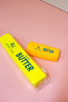 Small golden-yellow butter squishy beside a larger packaged yellow butter-stick toy on a pink surface.