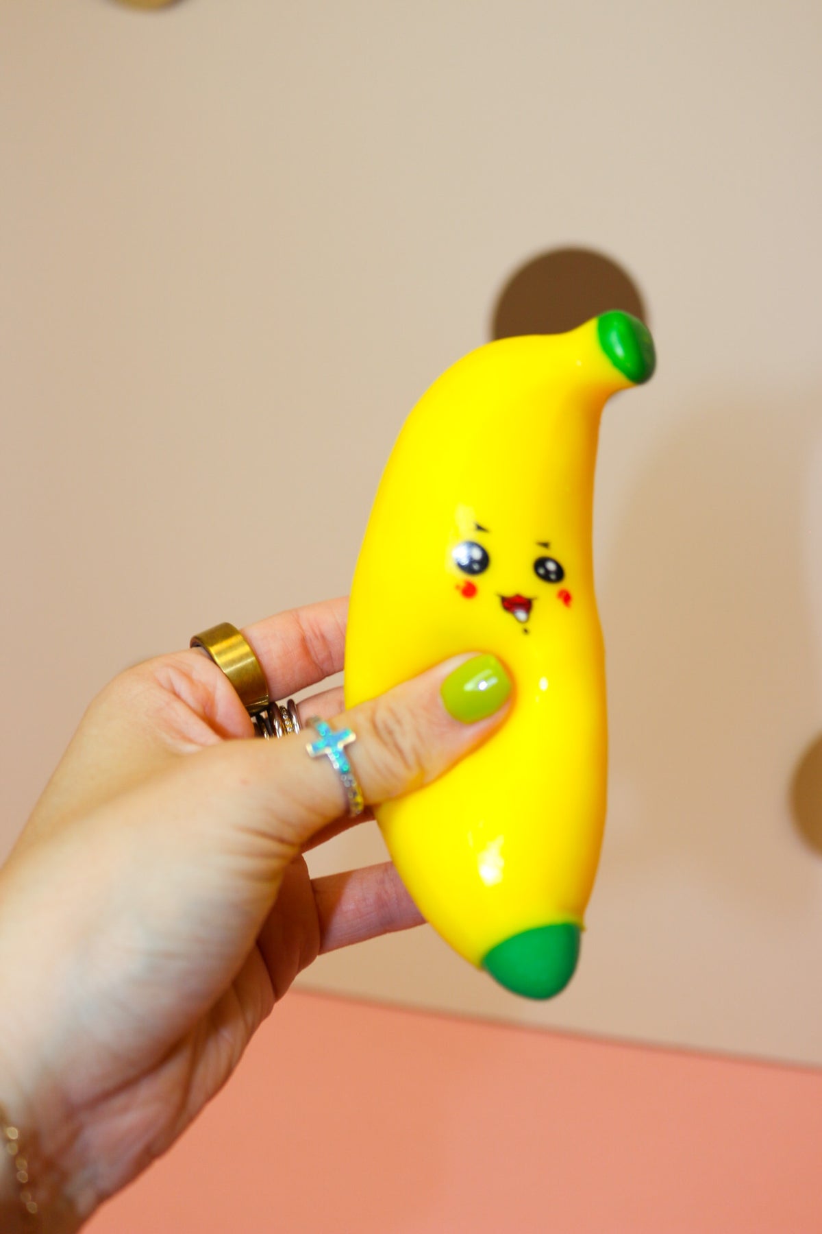 Hand squeezing a bright yellow banana squishy with green ends, cartoon eyes, rosy cheeks, and a smiling face.