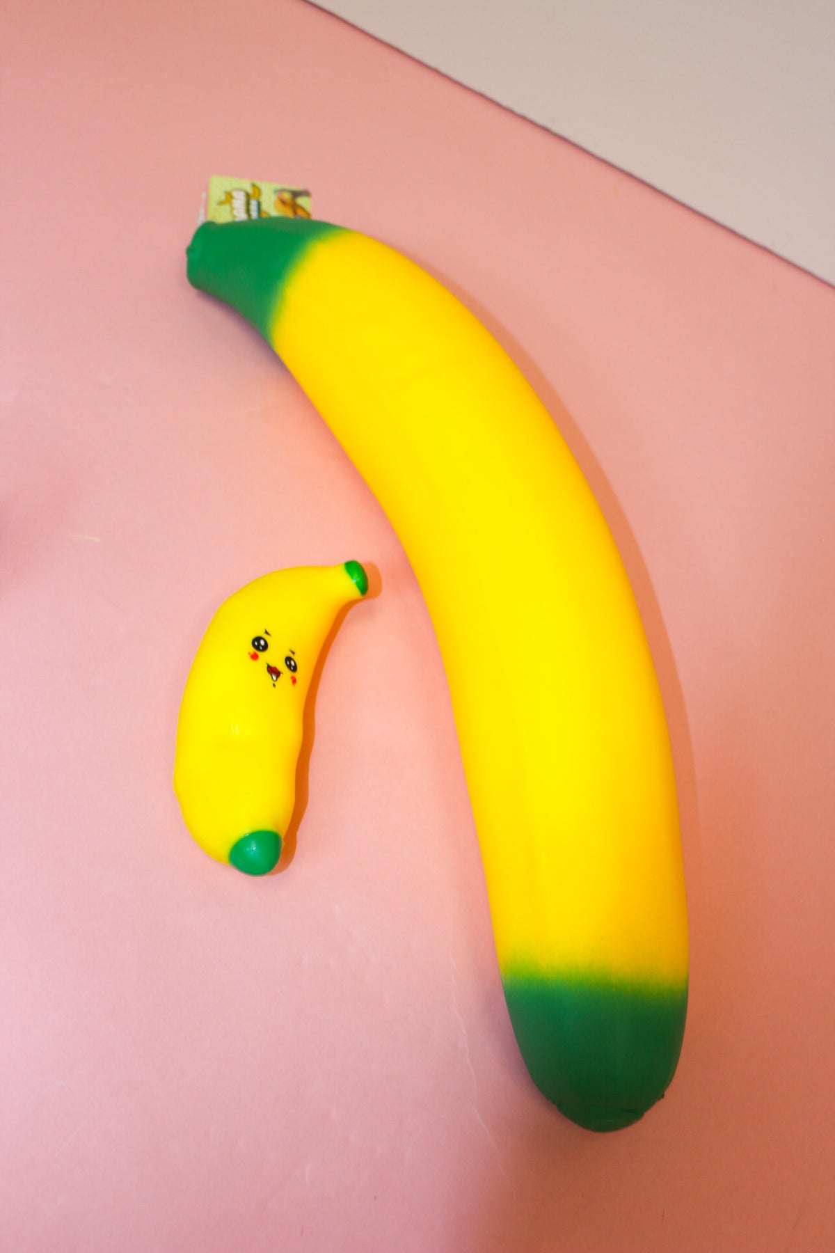 Small smiling yellow banana squishy with green ends beside a much larger banana-shaped item on a pink surface.