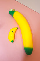 Small smiling yellow banana squishy with green ends beside a much larger banana-shaped item on a pink surface.