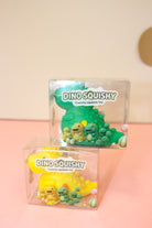 Green and yellow dinosaur squishy toys in clear Dino Squishy boxes stacked on a pink surface.