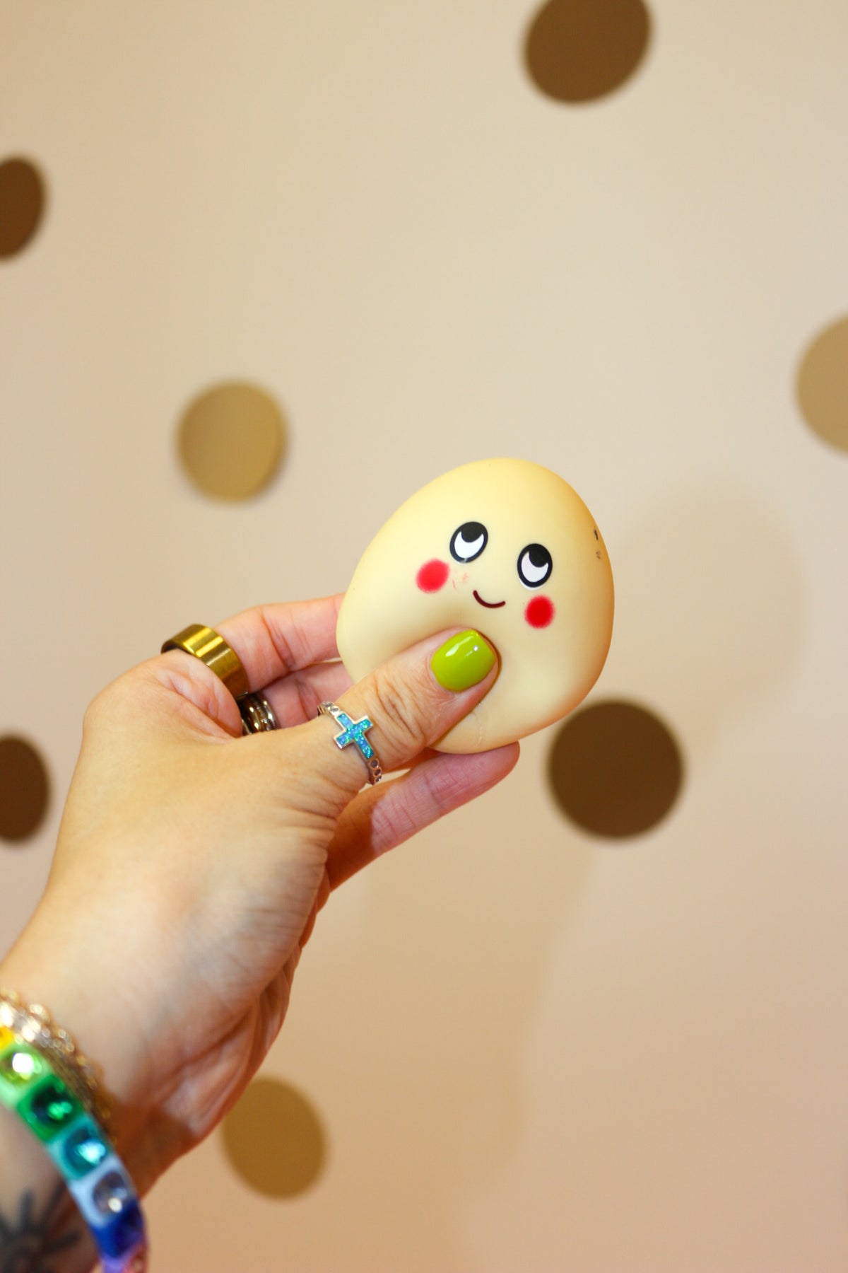 Hand squeezing a pale yellow egg-shaped squishy toy with cartoon eyes, red cheeks, and a smiling face.