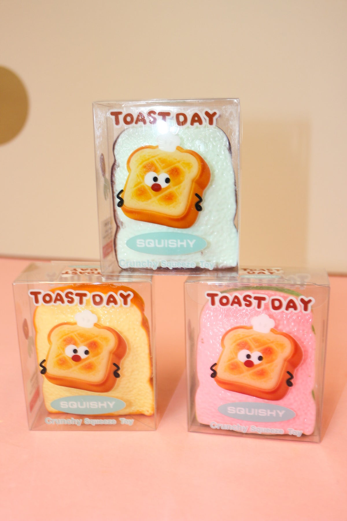 Three Toast Day squishy packages in pale blue, yellow, and pink stacked on a pink tabletop.